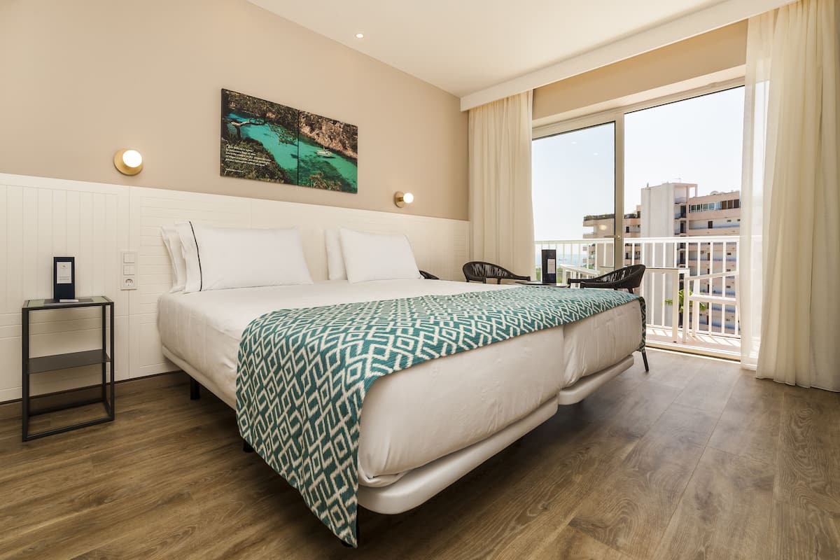 A hotel room with a large bed, a small side table, a wall-mounted light, and a balcony with a view of buildings.