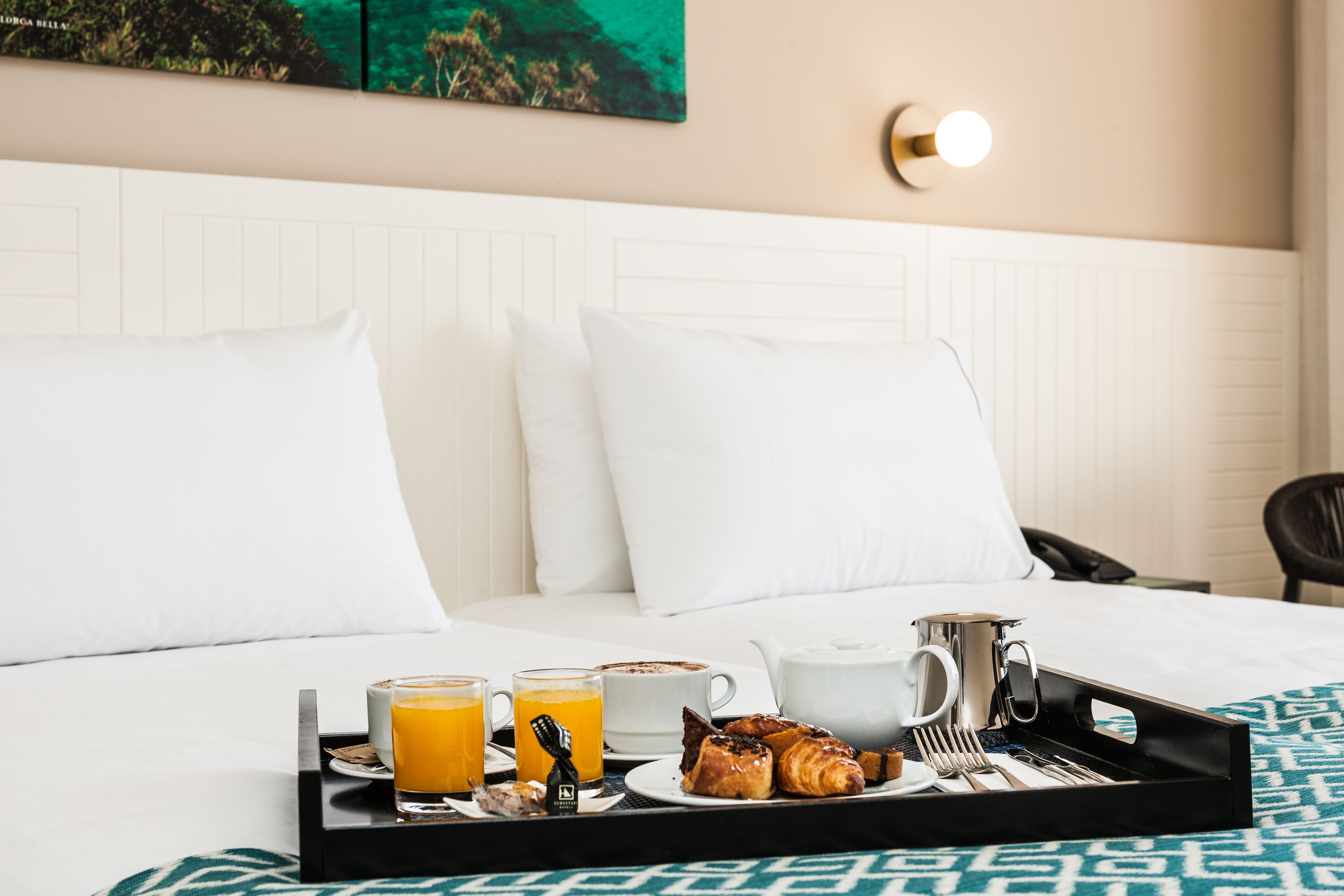 A neatly made bed with white linens, a tray with breakfast items including croissants, orange juice, and coffee, and a wall-mounted light fixture.