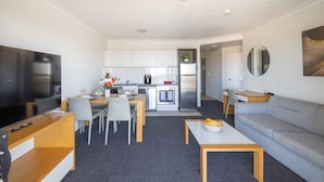 A modern living room with a flat-screen TV, a sofa, a dining table with chairs, and a kitchen area with white cabinets and appliances.