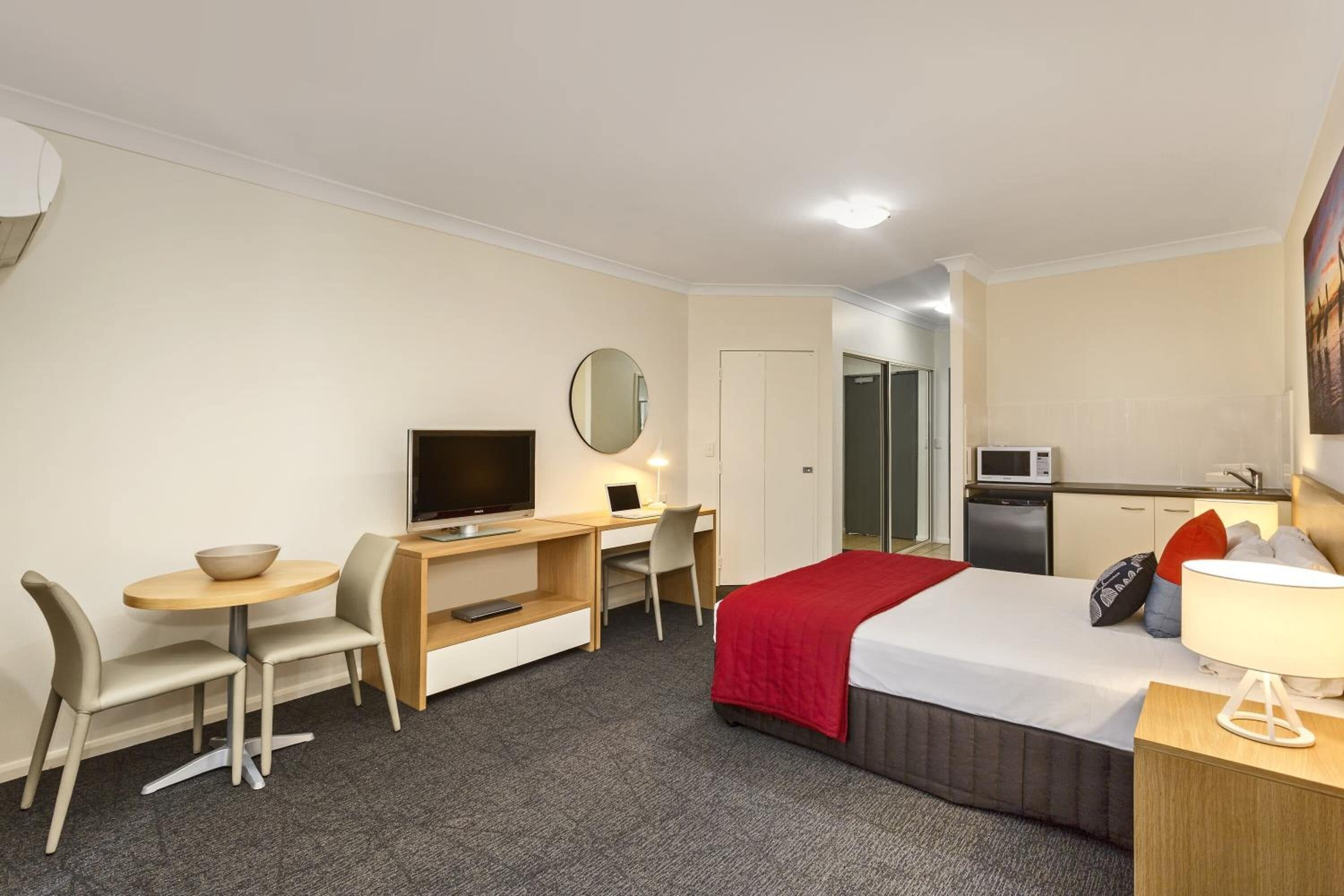 A hotel room with a bed, desk, chair, television, microwave, and a small kitchenette.