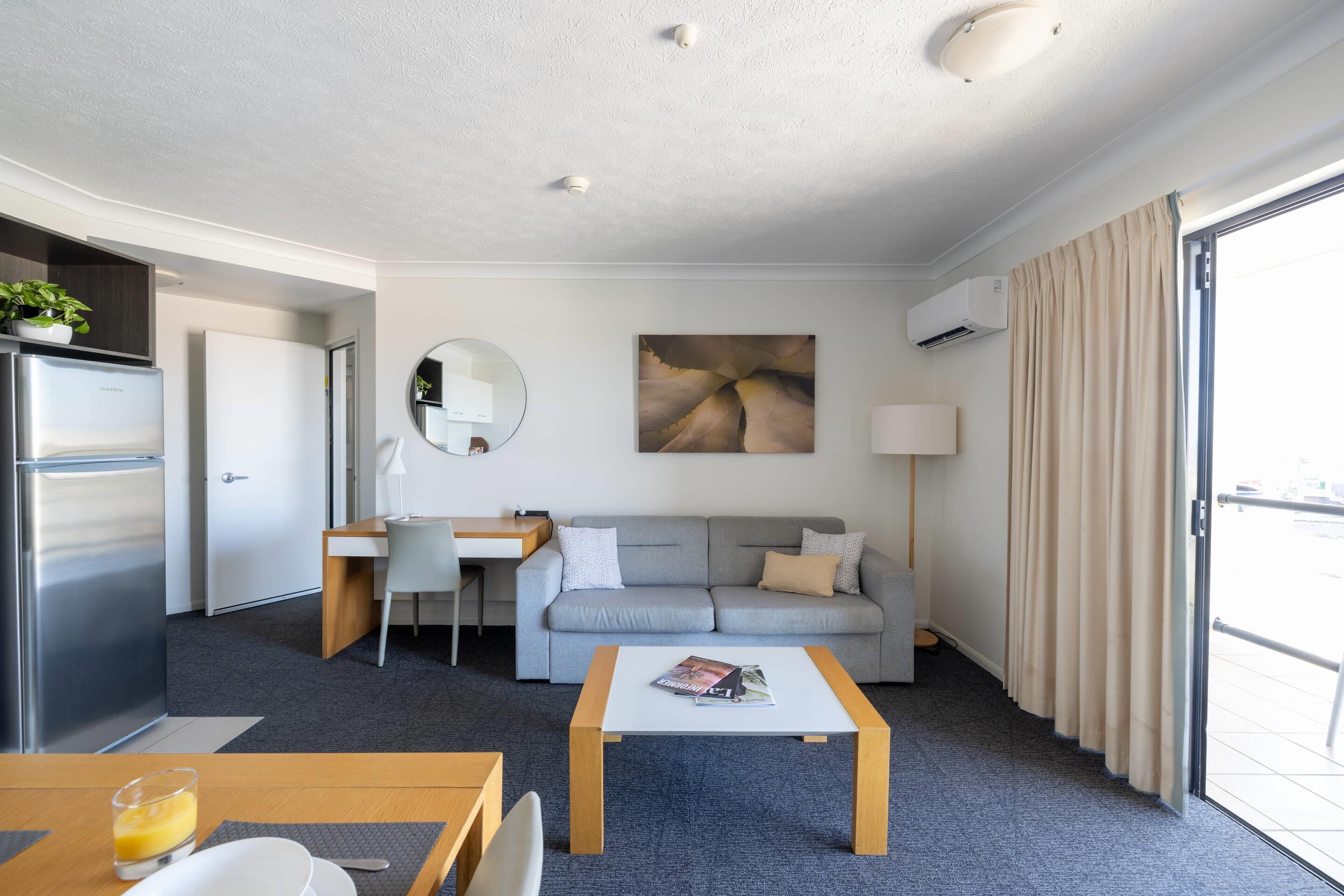 A modern hotel room with a kitchenette, a sofa, a dining table, and a large window with curtains.