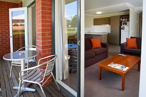 A balcony with a glass door, a metal table, and chairs. Inside, there is a living area with a sofa, a coffee table, and a kitchen area with cabinets and a microwave. - Quest Bendigo (Golden Square)