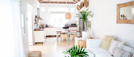 A spacious living area with a white sofa, a coffee table, and a dining table with chairs. There is a ceiling fan, a large plant, and a wooden ceiling.