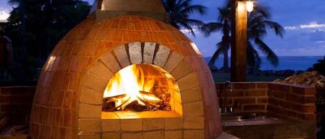 Un four à pizza en briques installé à l’extérieur, avec un feu qui crépite à l’intérieur, entouré d’une structure en bois offrant une vue sur l’océan et les palmiers en arrière-plan.