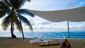 Eine Strandansicht mit Sonnensegel, Kissen, Feuerstelle und Esstisch, gedeckt für eine Mahlzeit.