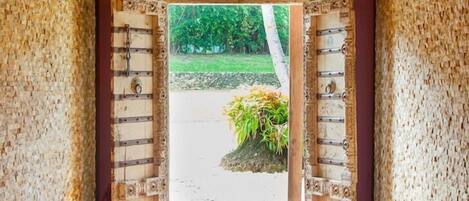 Une porte en bois ouverte, ornée de sculptures complexes, qui donne sur un espace extérieur avec un mur en pierre et une vue sur la verdure.