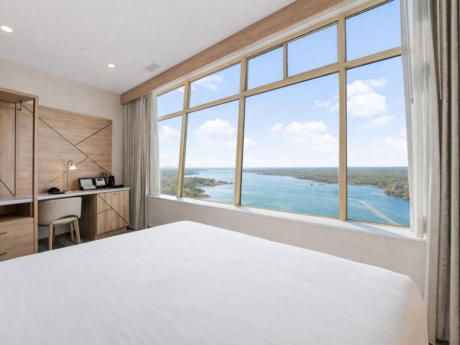 Ein Hotelzimmer mit einem großen Bett, einem Schreibtisch mit Stuhl und Blick auf ein Gewässer durch große Fenster.