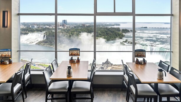 A dining area with large windows overlooking a waterfall and a cityscape. Tables are set with chairs and condiments.