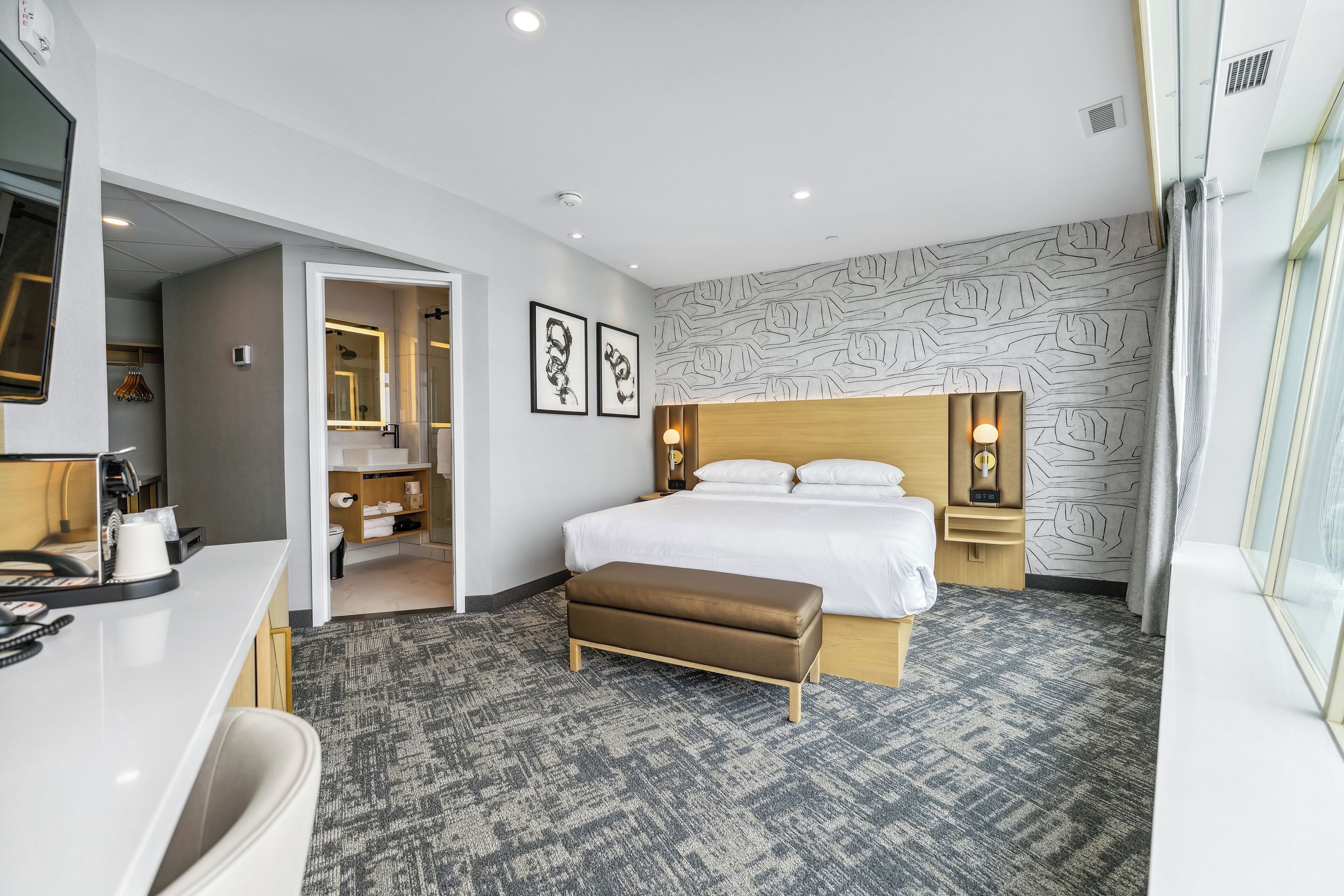 A hotel room with a bed, bedside tables, a desk, a chair, a bathroom, and a patterned wall.