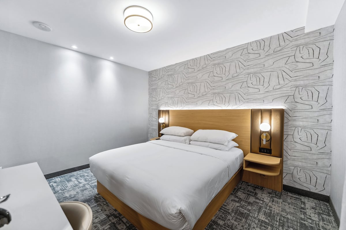 A modern hotel room with a neatly made bed, a bedside table with a lamp, and a patterned wall.
