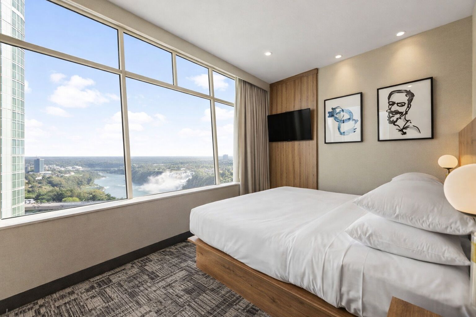 A modern hotel room with a large window offering a city view, a bed with white linens, a mounted TV, and two framed artworks on the wall.