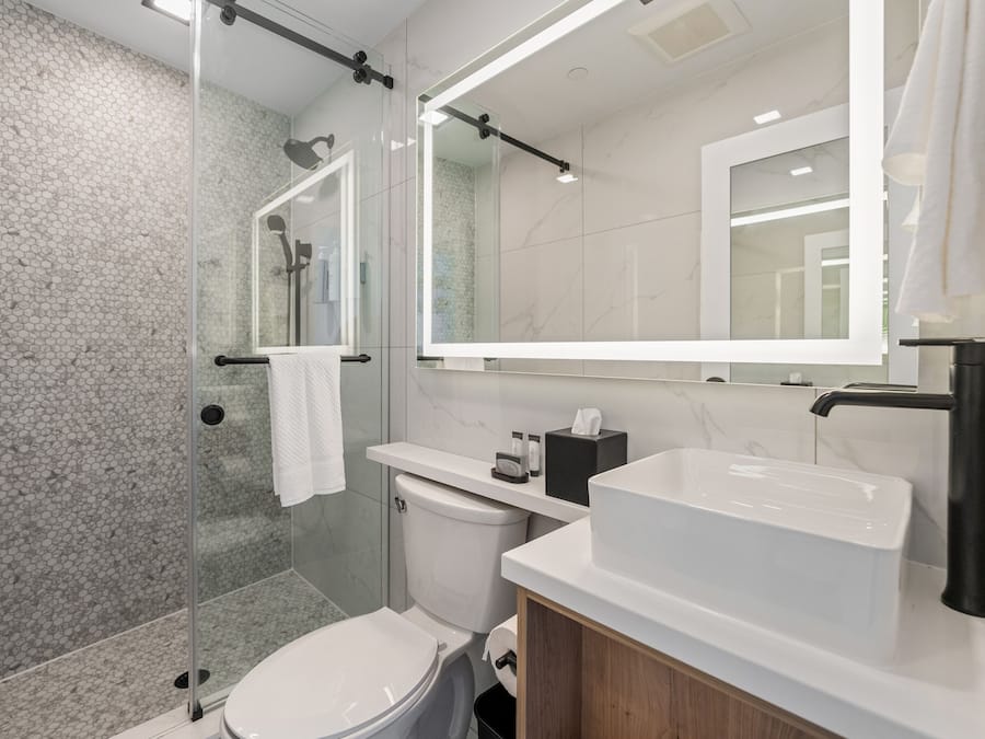 Un baño moderno con cabina de ducha de vidrio, inodoro blanco, tocador con un espejo grande y lavabo blanco.