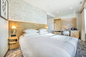 Une chambre d’hôtel avec un grand lit, un bureau et une tête de lit ornée d’un motif de feuilles. On y trouve des lampes fixées au mur et une petite étagère avec une horloge.