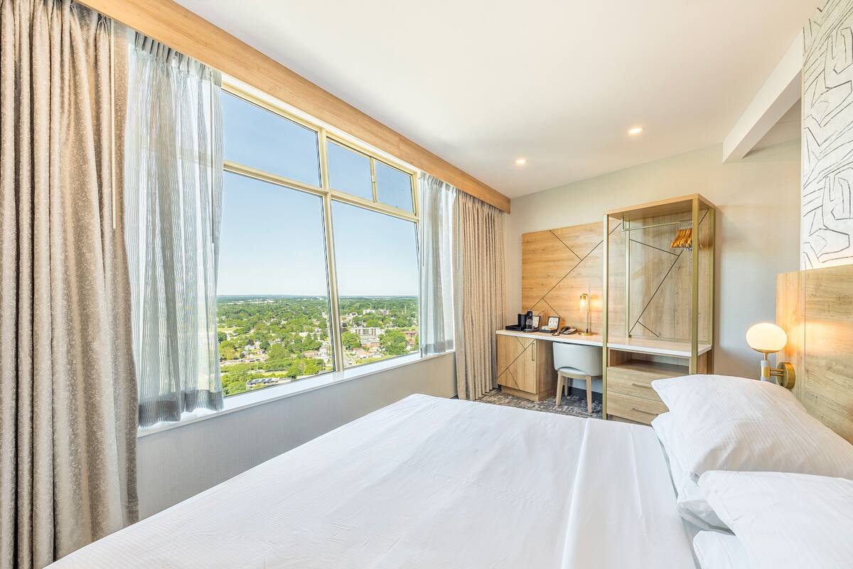 A hotel room with a large window, a desk, a wardrobe, and a bed.