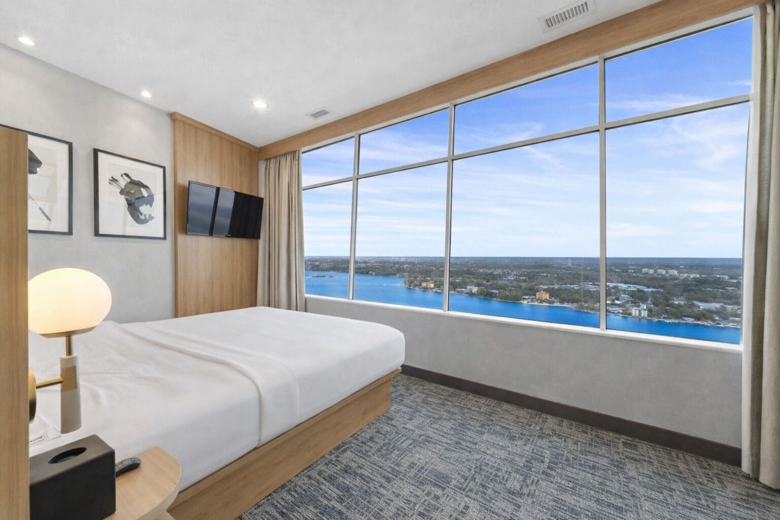 A hotel room with a large window, a bed, a TV, and framed artwork on the wall.