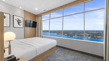 A hotel room with a large window, a bed, a TV, and framed artwork on the wall.