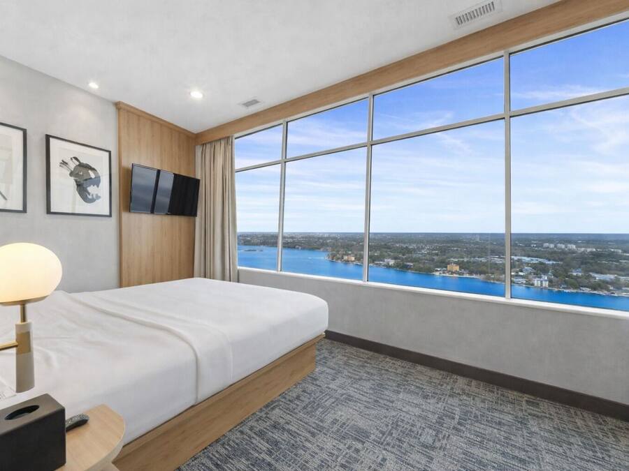 A hotel room with a large window, a bed, a TV, and framed artwork on the wall.
