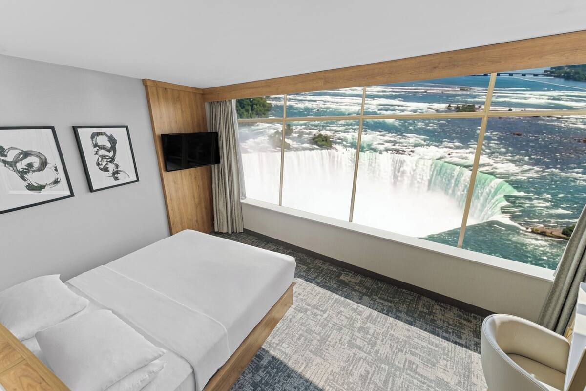 A hotel room with a large window overlooking a waterfall, a bed, a TV, and artwork on the wall.