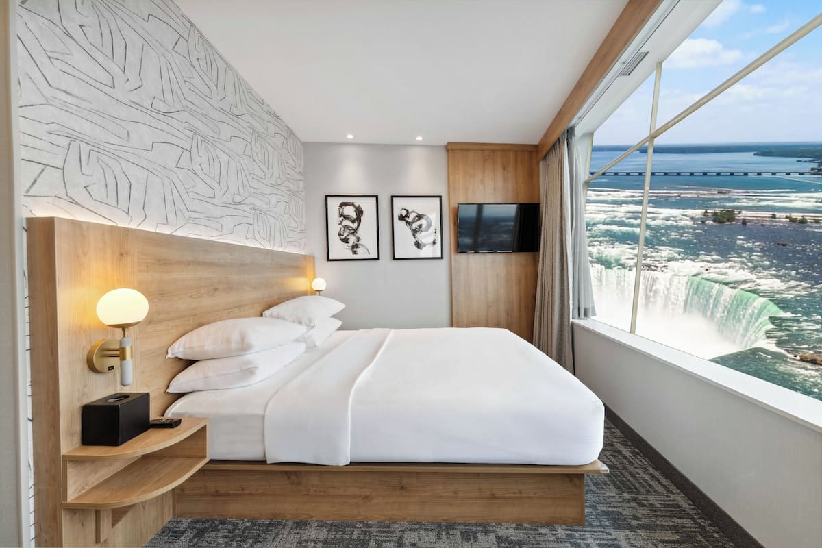 A hotel room with a large bed, a nightstand with a lamp, two framed pictures, a TV, and a view of a waterfall.