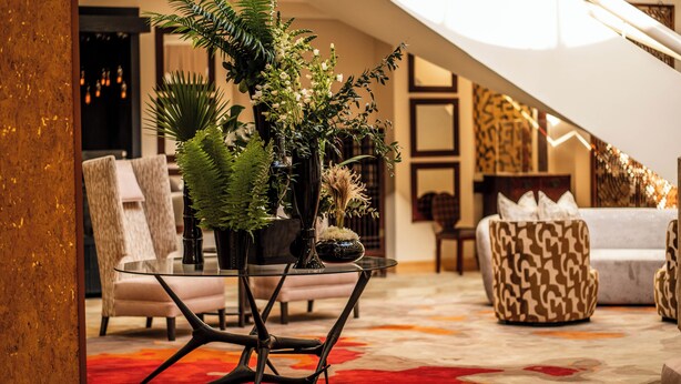 A modern living room with a glass coffee table, patterned sofa, and decorative plants.