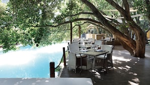 An outdoor dining area with tables and chairs under large trees, overlooking a pool. - Saxon Hotel, Villas and Spa (Sandton)