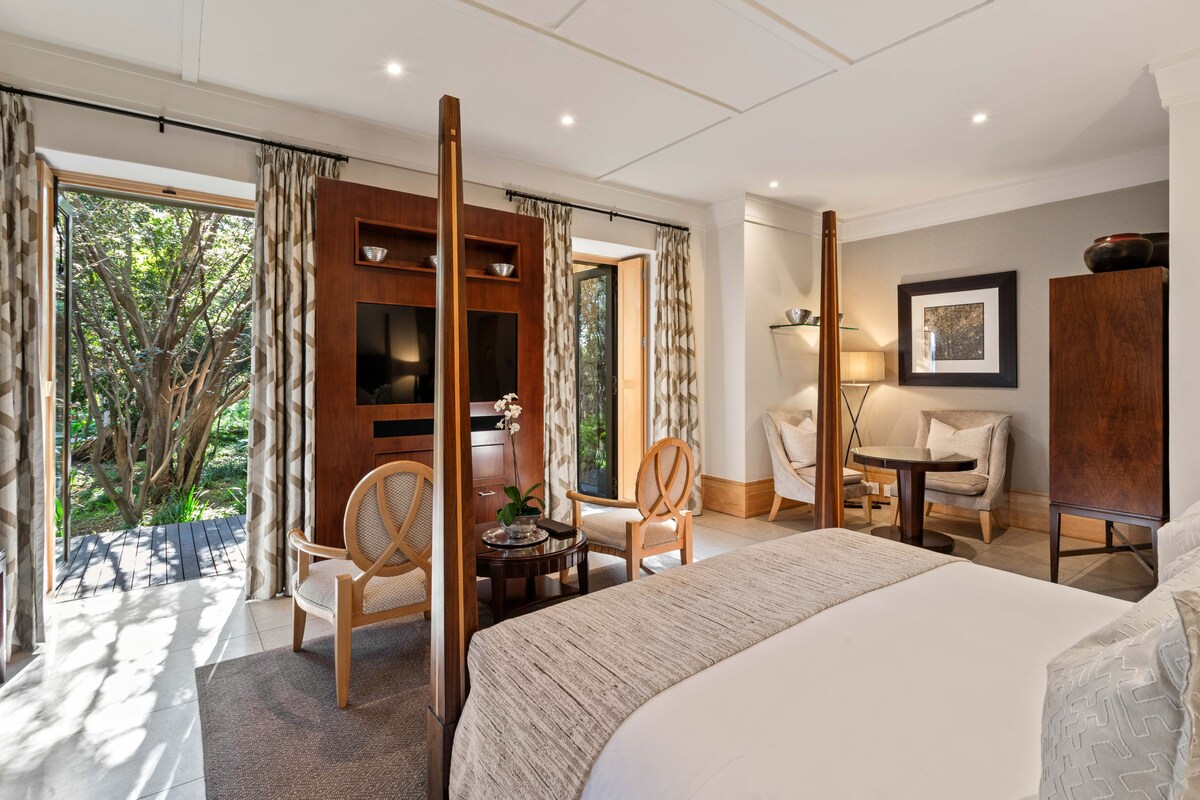 Ein Hotelzimmer mit einem großen Bett, zwei Sesseln, einem kleinen Tisch und Blick ins Freie.