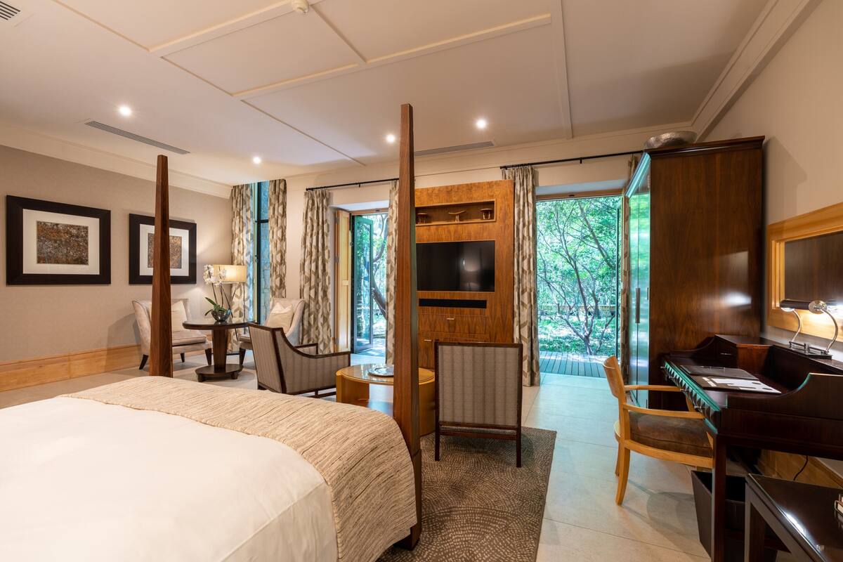 Habitación de hotel con una cama grande, un escritorio con un piano, televisión y vistas a zonas verdes por las ventanas.