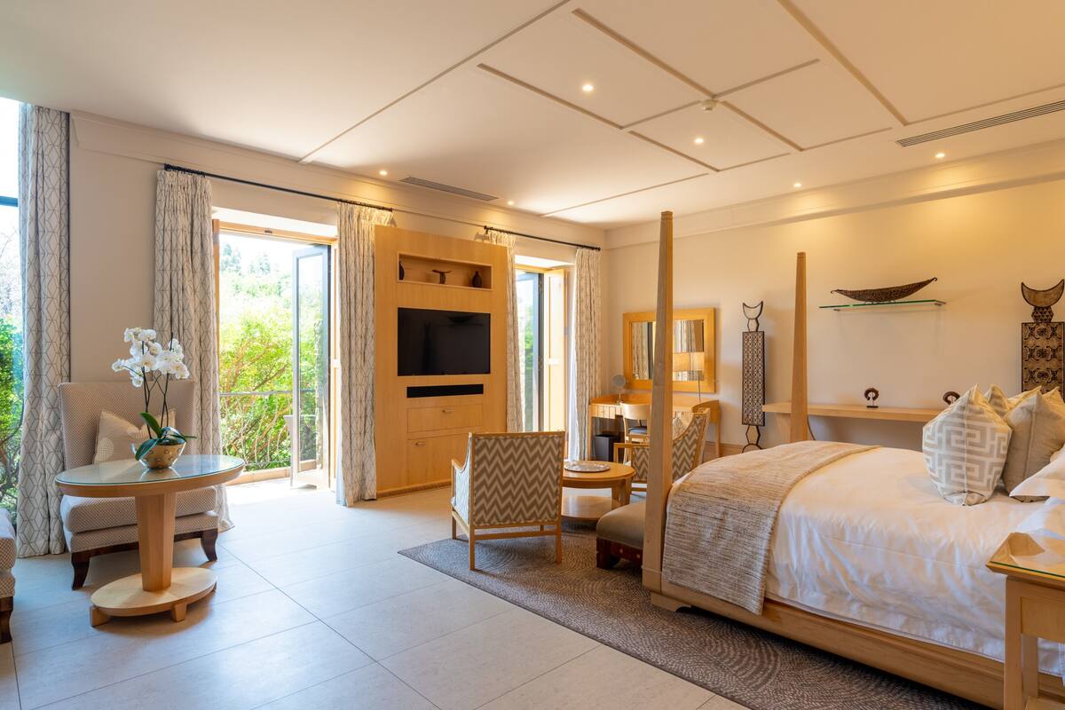 Habitación de hotel moderna con una cama grande, un televisor, un área de estar con una mesa y sillas, y un balcón con cortinas.