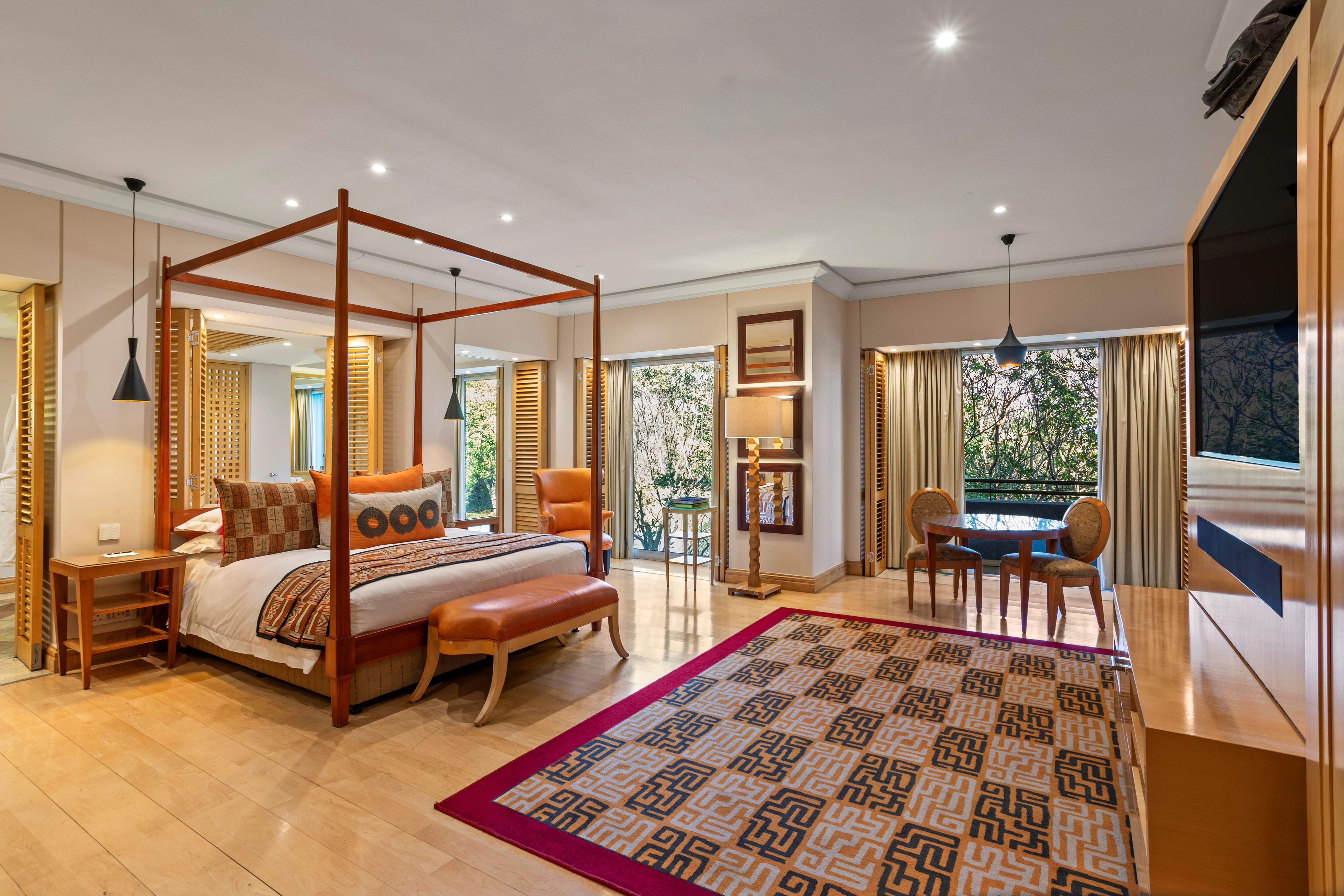 A spacious bedroom with a four-poster bed, a sitting area with a table and chairs, and a large window offering a view of greenery.
