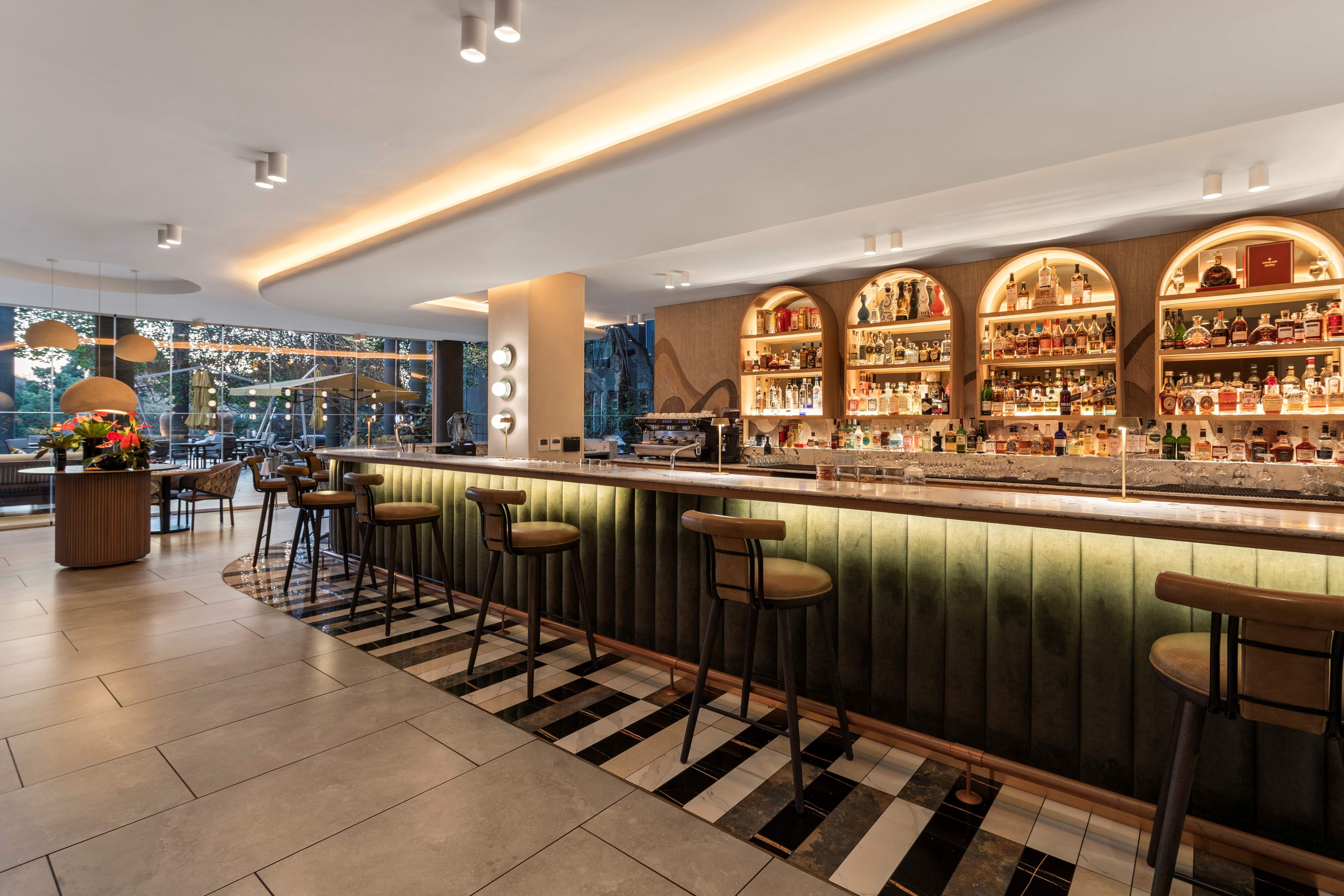 Een bar met een zwart-wit schaakbordvloer, barstoelen en een goed verlichte plank met flessen.