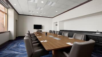 Una sala de conferencias con una larga mesa, sillas de cuero, un televisor de pantalla plana y una ventana con persianas.