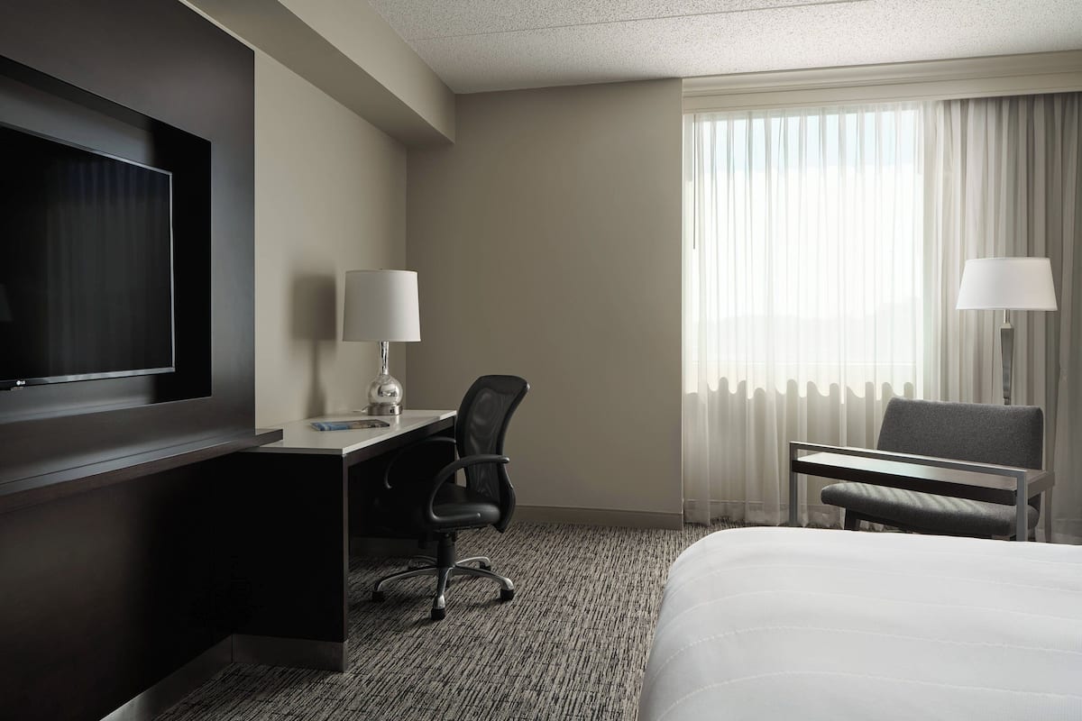 A hotel room with a bed, a desk with a chair, a TV, and a window with curtains.