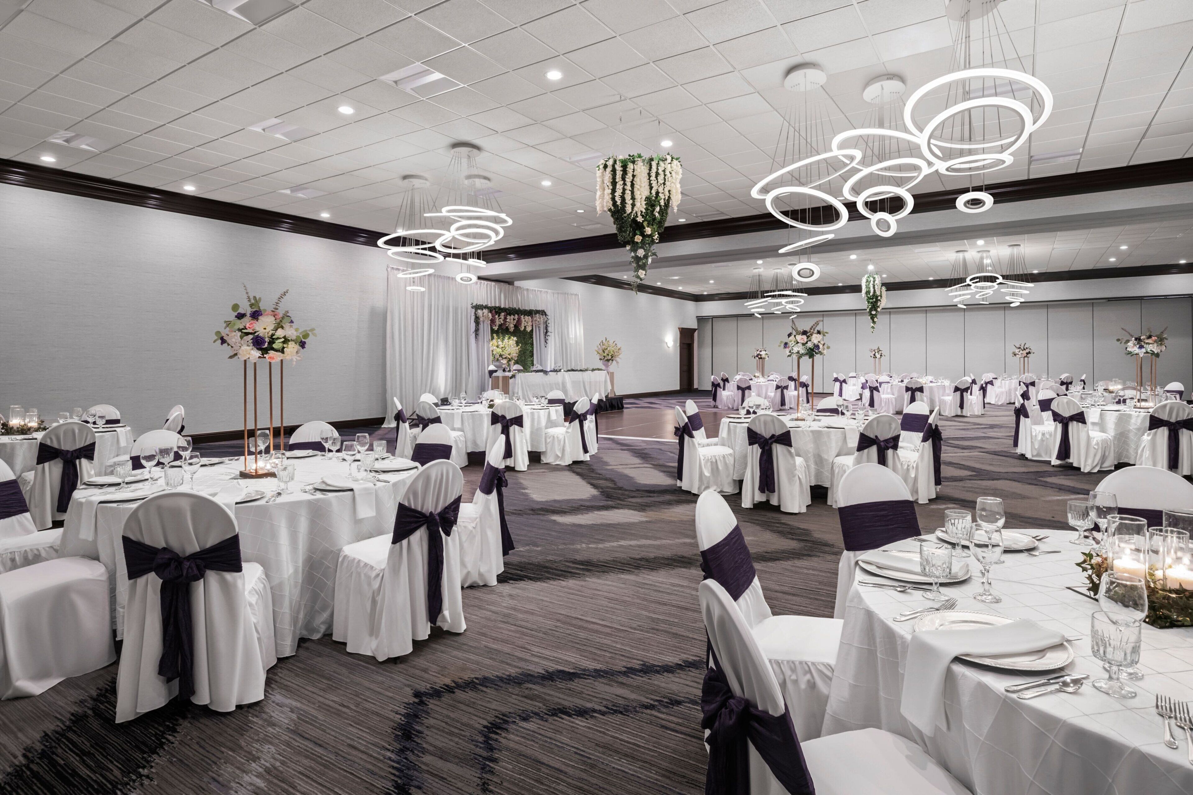 Un salón de banquetes con mesas redondas, manteles blancos y sillas con cintas oscuras.