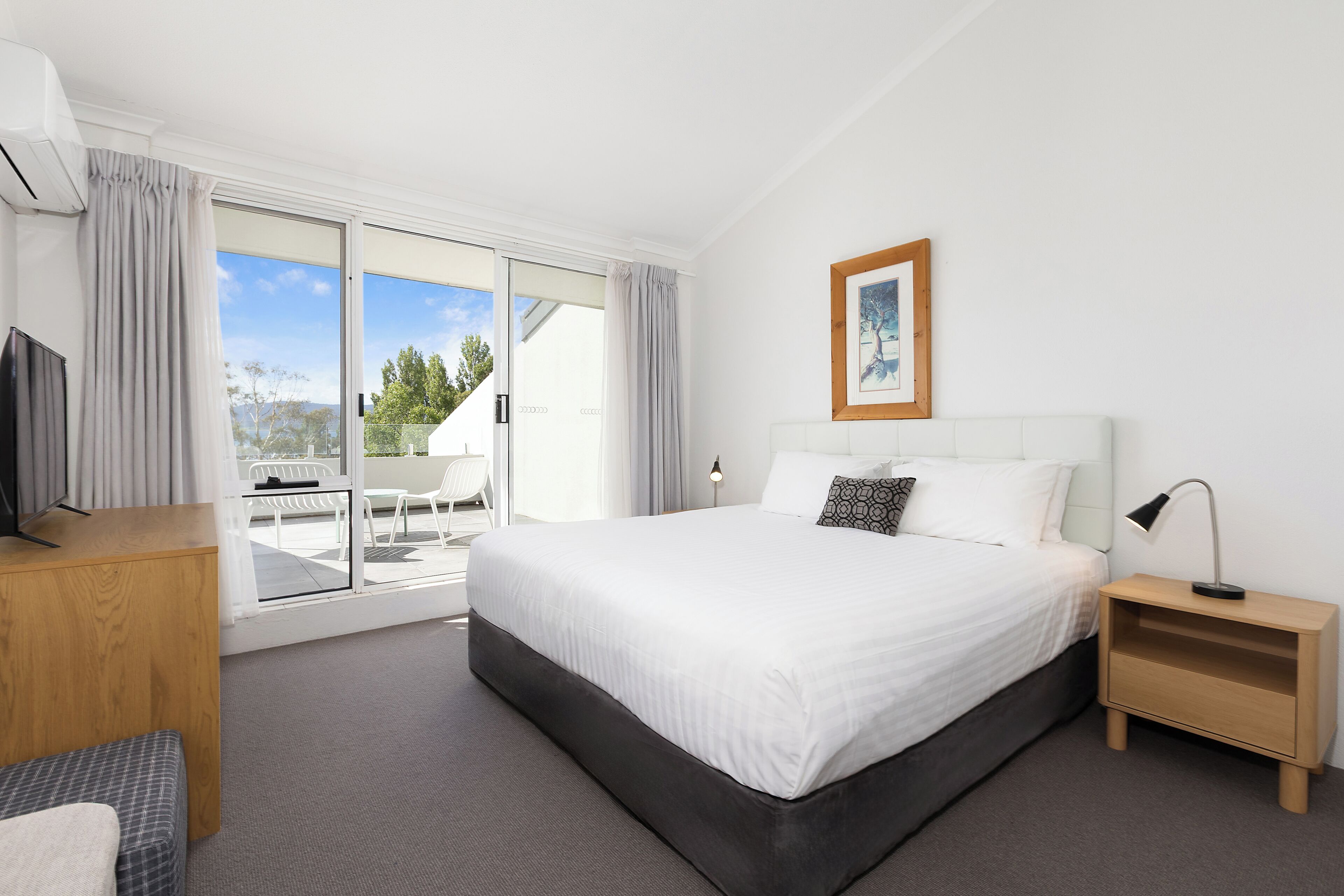 A hotel room with a bed, a desk, a chair, a TV, a nightstand, and a window with curtains.