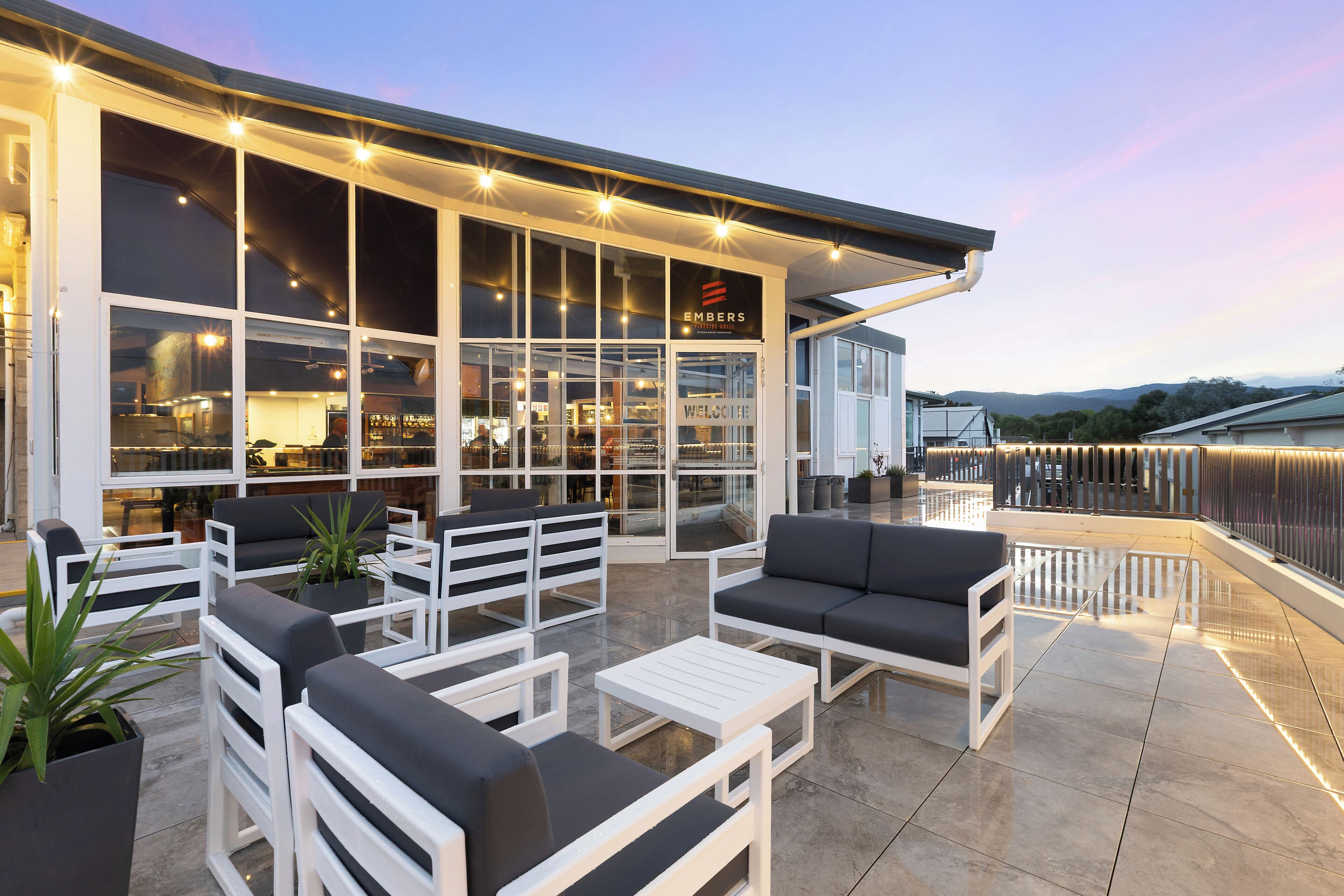 An outdoor seating area with white furniture and potted plants, a modern building with large windows, and a sign reading 'EMBERS.'