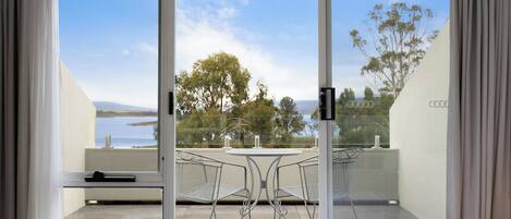 A balcony with a table and chairs, a bed, and a view of the sea and trees.