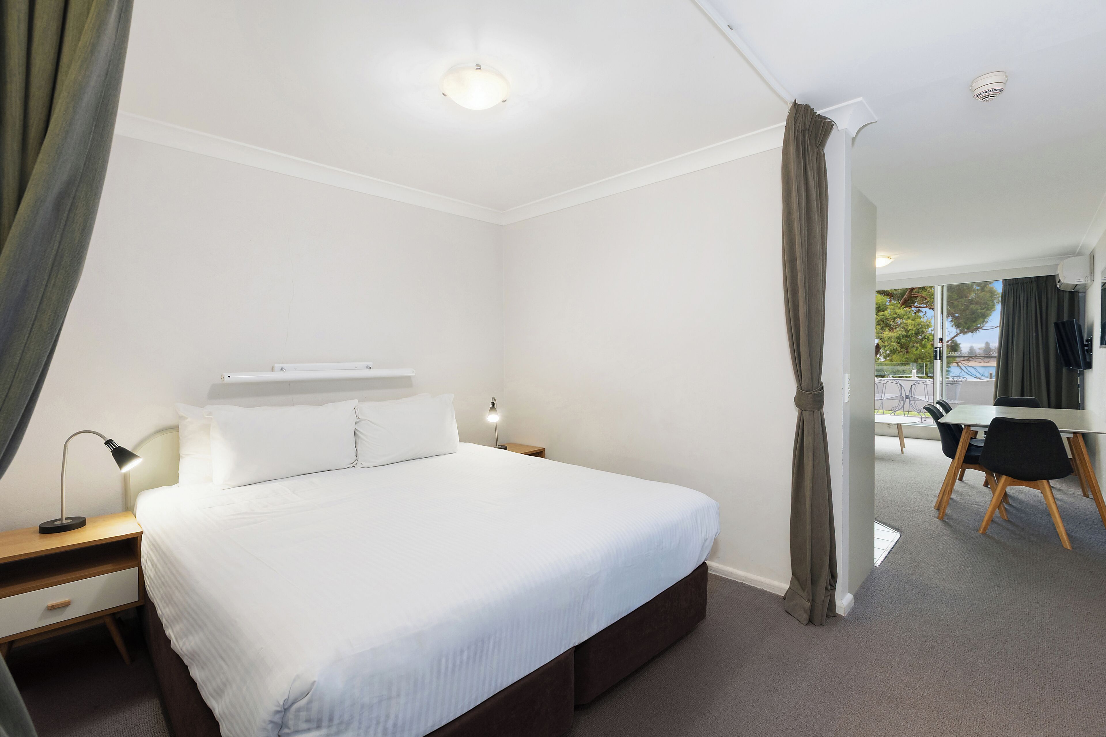 A hotel room with a bed, bedside table, lamp, and a view of the outside.