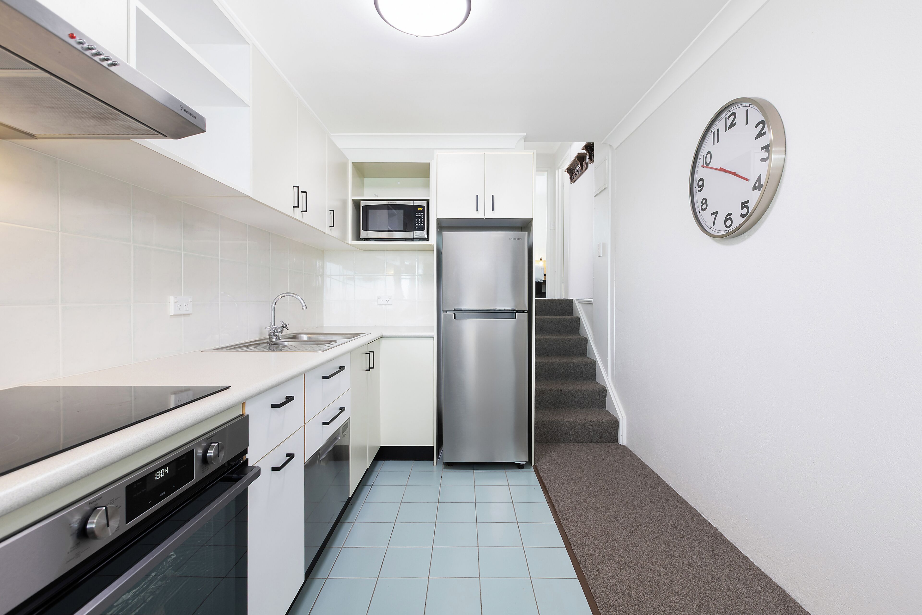 Una cocina moderna con refrigerador de acero inoxidable, microondas y horno, gabinetes blancos y un reloj en la pared.