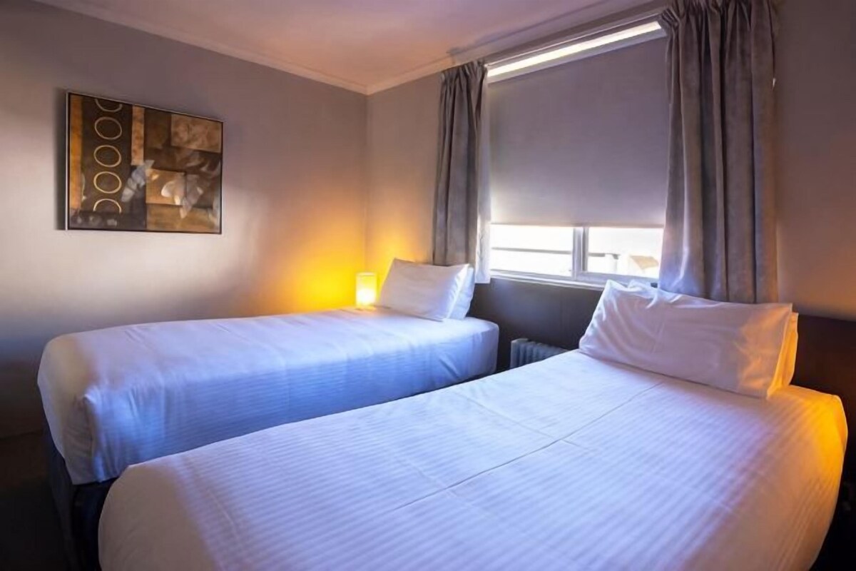 Habitación de hotel con dos camas, una ventana con cortinas, un cuadro en la pared y una lámpara en la mesita de noche.