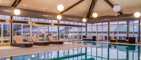 A modern indoor swimming pool with clear glass walls, lounge chairs, and ceiling lights.