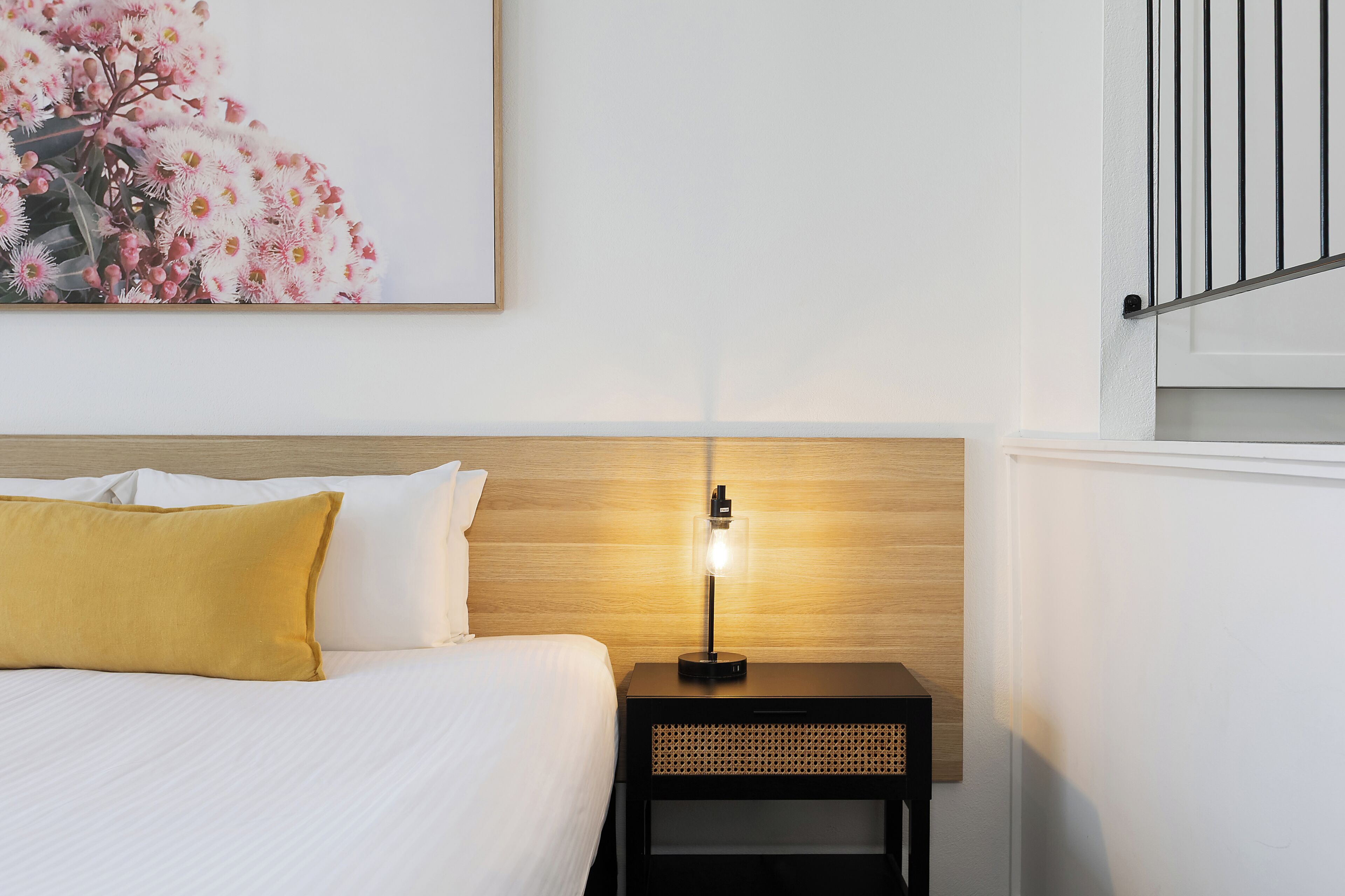 A neatly made bed with white and yellow pillows, a bedside table with a lamp, and a wall-mounted picture of flowers.
