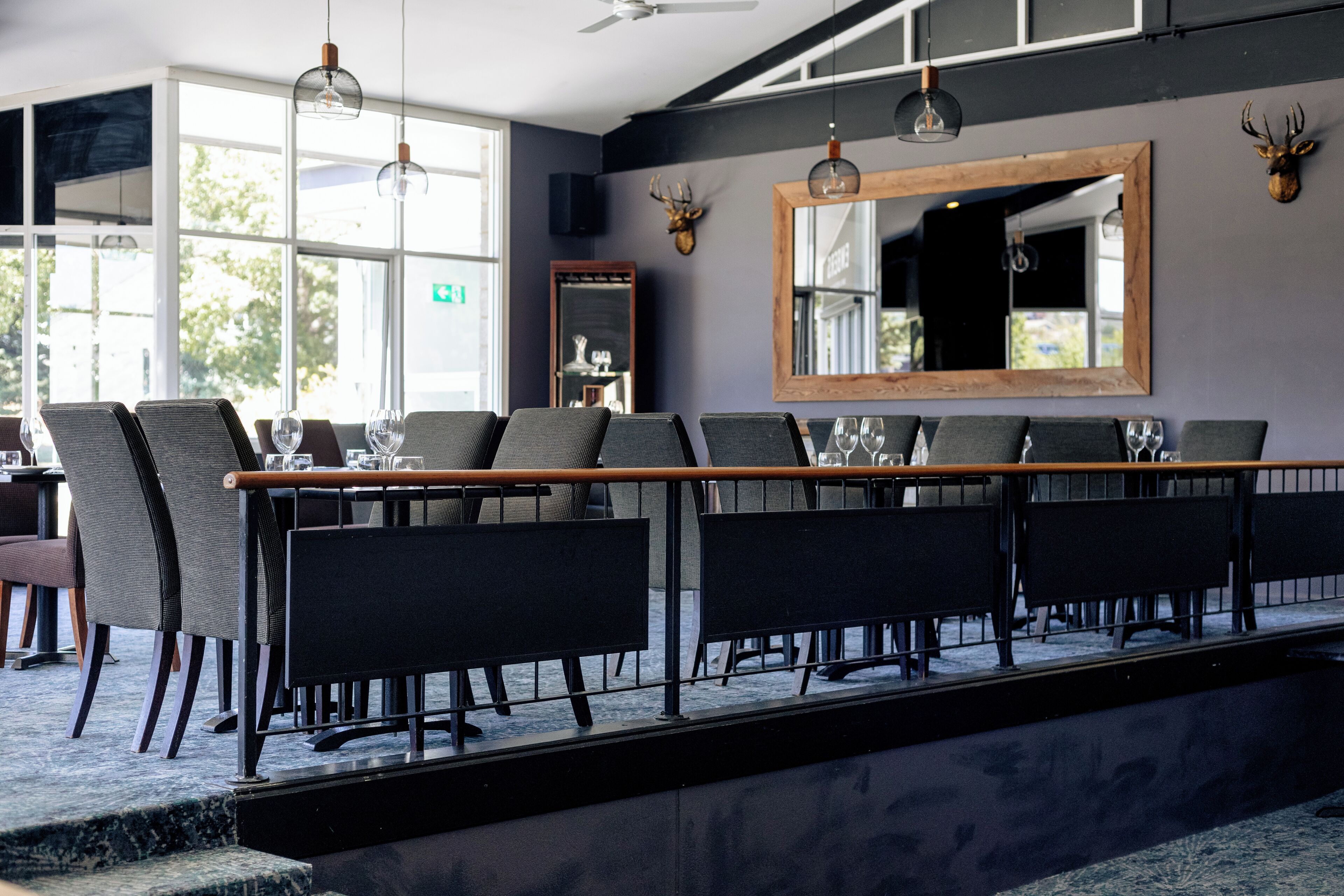 En restaurang med ett långt bord, stolar och en räcke. Det finns stora fönster, en takfläkt och en spegel på väggen.