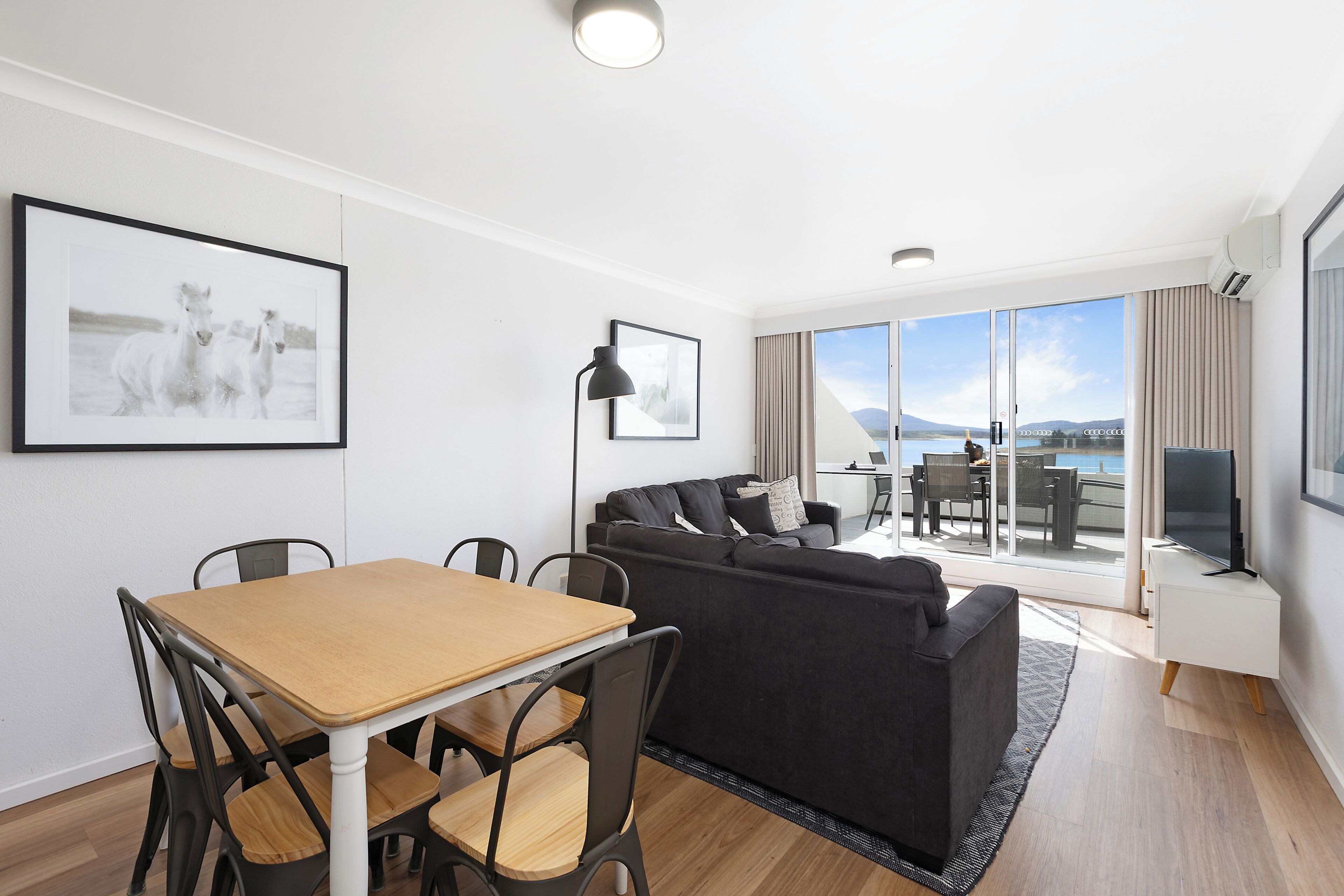 A modern living area with a dining table, black sofa, and large windows offering a view of the outdoors.