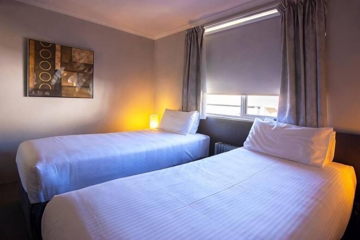 A hotel room with two beds, a window with curtains, a painting on the wall, and a lamp on the nightstand.