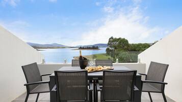 Une table avec quatre chaises, une bouteille de champagne et une assiette de nourriture, le tout sur une terrasse donnant sur un lac, avec des montagnes en arrière-plan.