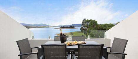 A table with four chairs, a bottle of champagne, and a plate of food on a terrace overlooking a lake with mountains in the distance.