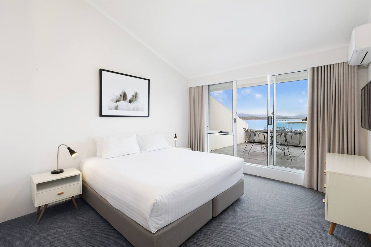 A hotel room with a bed, a nightstand, a desk, a chair, and a large sliding glass door leading to a balcony with a view.