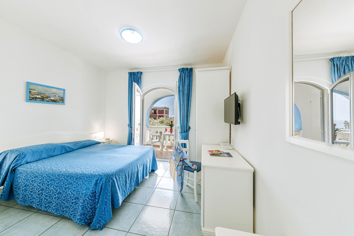 Quarto com cama, escrivaninha, cadeira, televisão e janela com cortinas.