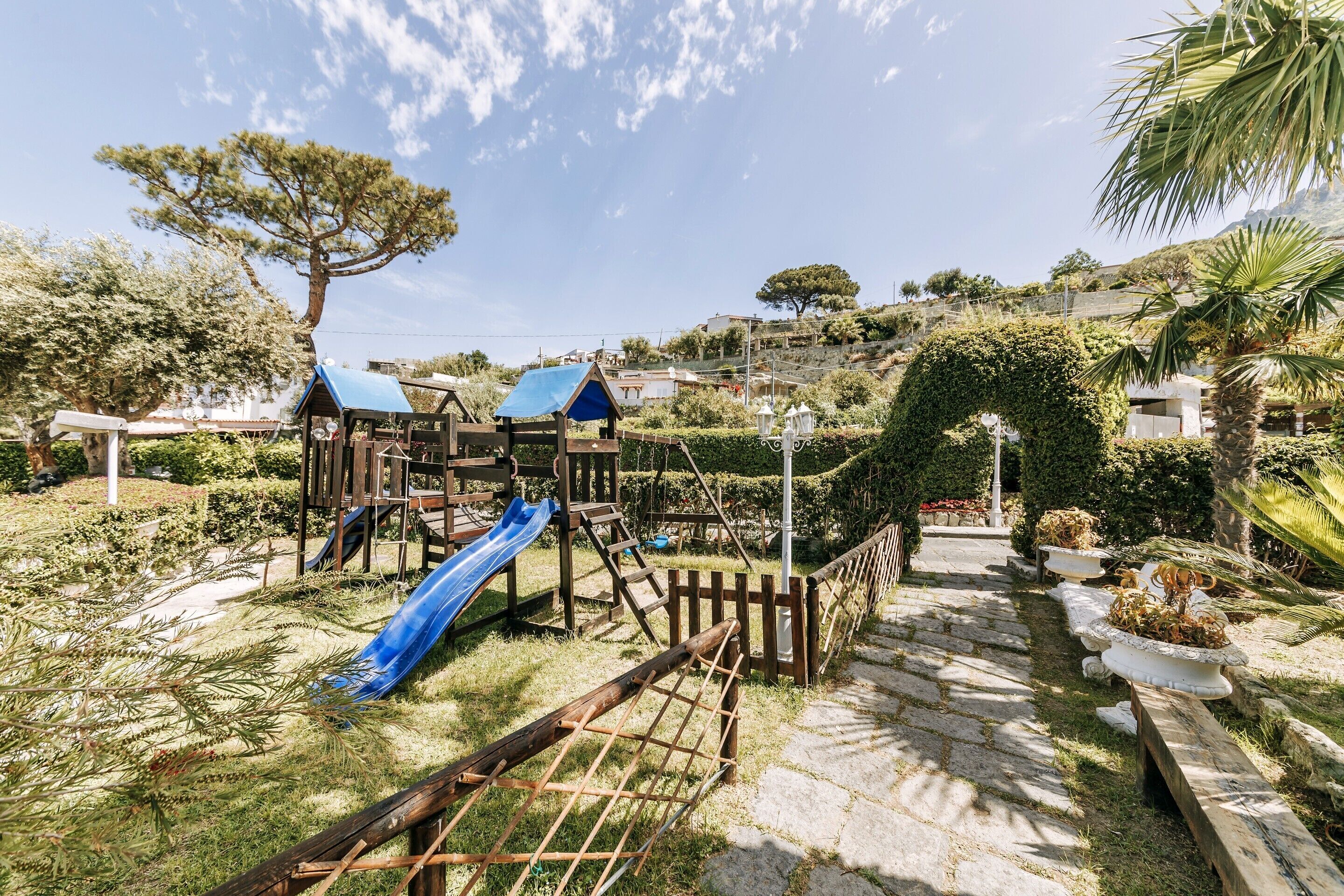Un parque infantil con un tobogán y estructuras para escalar, rodeado de jardines cuidados y un camino de piedra.