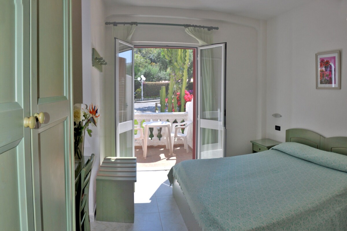 Quarto com cama, uma mesinha e uma cadeira em frente a uma porta de correr de vidro.